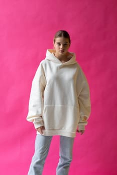 Beautiful blonde woman in a white hoodie and blue jeans posing on a pink background. High quality photo