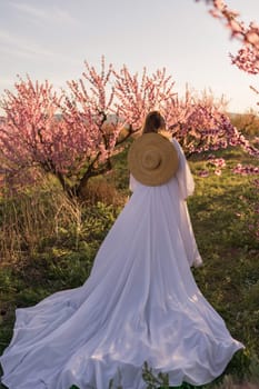 Woman blooming peach orchard. Against the backdrop of a picturesque peach orchard, a woman in a long white dress and hat enjoys a peaceful walk in the park, surrounded by the beauty of nature