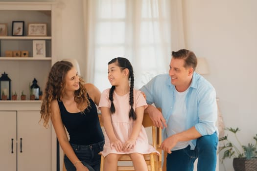 Happy family portrait with lovely little girl smiling and looking at camera, lovely and cheerful parent and their daughter sitting together in living room at home with warm daylight. Synchronos