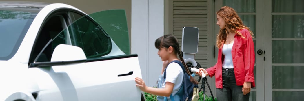 Happy little young girl learn about eco-friendly and energy sustainability as she help her mother recharge electric vehicle from home EV charging station. EV car and modern family. Panorama Synchronos