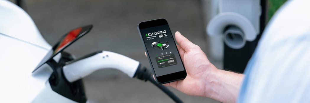 Modern eco-friendly man recharge electric vehicle from EV charging station. EV technology utilization for tracking energy usage to optimize battery charging display on smartphone. Panorama Synchronos