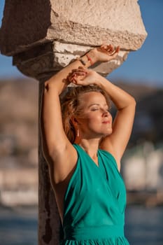 Happy blonde in a long mint dress posing against the backdrop of the sea in an old building with columns. Girl in nature against the blue sky