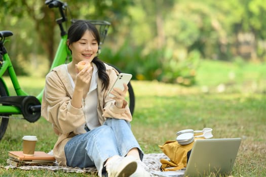 Cheerful young female student sitting on grass in the campus and using mobile phone.0