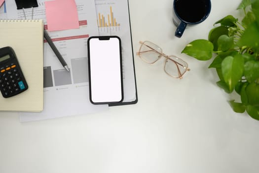 Top view smartphone with empty screen, glasses and business reports on white table.