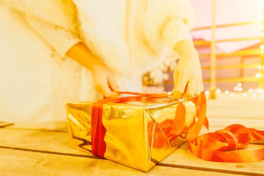 A woman in a white dress is holding a gold box with a red ribbon. She is wearing a crown on her head. The scene takes place in a room with a door and a window. The woman appears to be opening the gift box.