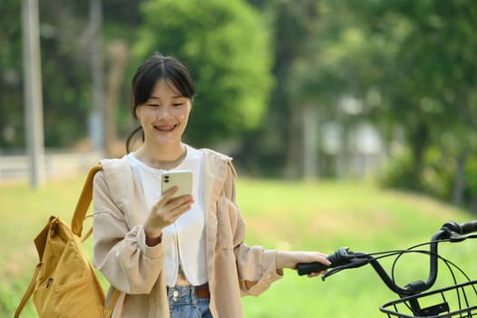 Smiling young woman with backpack standing near bicycle in city park and chatting on mobile phone.