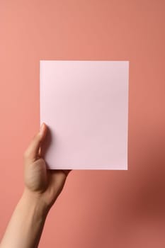 A hand holding a blank pink square paper against a pink background.