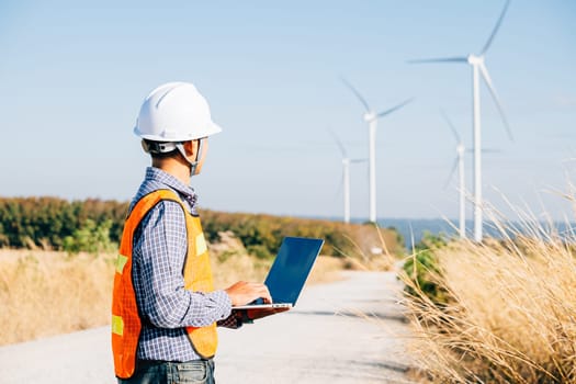 Engineer with laptop by windmills working on efficiency. Service technician ensures turbine performance. Landscape development innovation for windmill service and quality confidently managed.