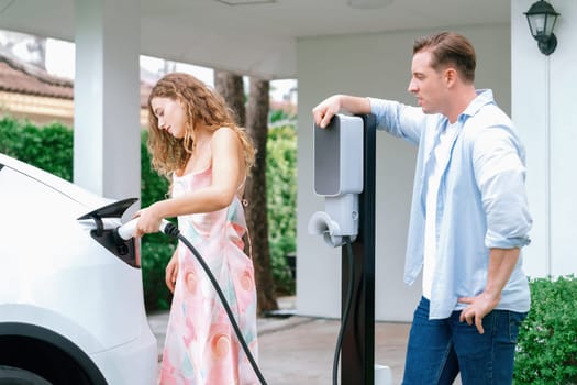 Happy and lovely couple with eco-friendly conscious recharging electric vehicle from EV home charging station. EV car technology utilized for residential home to future sustainability. Synchronos