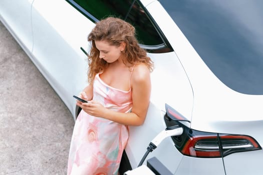 Aerial top view eco-friendly woman recharge electric vehicle from EV charging station, using EV technology utilization for tracking energy usage to optimize battery charging on smartphone. Synchronos