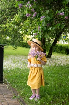 A little girl in a yellow dress and straw hat wearing a bouquet of lilacs. A walk in a spring park, blossoming lilacs