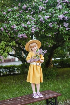 A little girl in a yellow dress and straw hat wearing a bouquet of lilacs. A walk in a spring park, blossoming lilacs