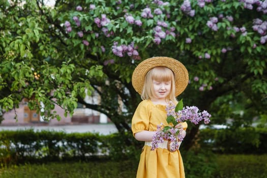 A little girl in a yellow dress and straw hat wearing a bouquet of lilacs. A walk in a spring park, blossoming lilacs