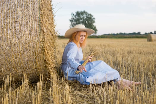 A red-haired woman in a hat and a blue dress walks in a field with haystacks