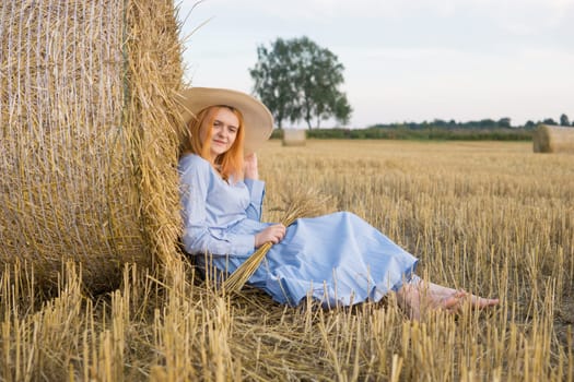 A red-haired woman in a hat and a blue dress walks in a field with haystacks