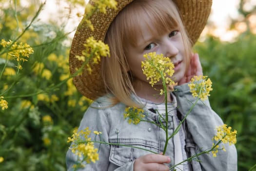 Blonde girl in a field with yellow flowers. A girl in a straw hat is picking flowers in a field. A field with rapeseed