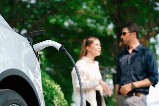 Young couple recharge electric car's battery from charging station in outdoor green city park in springtime. Rechargeable EV car for sustainable environmental friendly urban travel lifestyle.Expedient