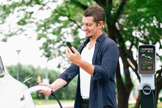 Man using smartphone online banking application to pay for electric car battery charging from EV charging station during vacation holiday road trip at national park or summer forest. Exalt