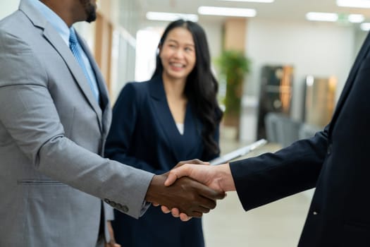 Closeup of business hands shaking between businessman and professional male leader while smart businesswoman stand near at modern office corridor. Represented unity, corporate, teamwork. Ornamented.