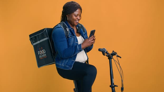 Smiling black woman texting coworkers with phone on bicycle before cycling for food delivery service job. African american lady checking mobile app for packages ready for delivering.