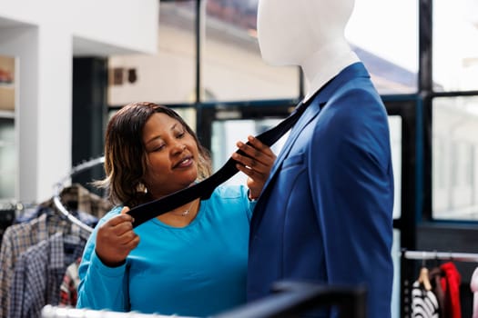 Stylish client looking at mannequin wearing formal tie, analyzing fabruc in modern boutique. African american customer shopping for trendy casual wear, clothing store full with fashionable merchandise