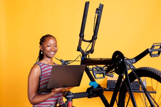 Female cyclist of african american ethnicity fixes damaged bike using toolkit and laptop to research solutions. Using minicomputer and professional equipment, black woman is repairing broken bicycle.