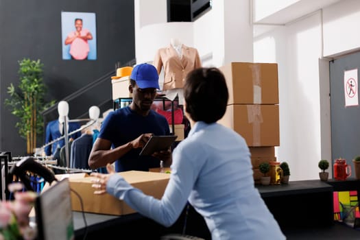 Store worker helping courier with packages, carrying cardboard boxes in modern boutique. African american deliveryman discussing shipping details with employee in shopping centre. Fashion concept