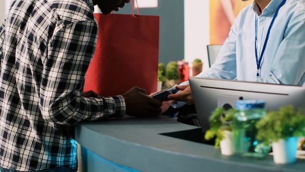 Employee giving paper bag to client after paying for fashionable merchandise in clothing store. African american shopaholic man purchasing stylish clothes and trendy accessories in modern boutique