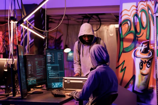 Hacker in hood showing black box with hacking technology to break into server database. Young criminal opening case with equipment for online cyberattack in abandoned warehouse