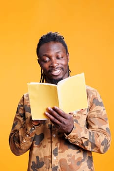 Intellectual african american man relaxing reading novel book, enjoying learning new information standing in studio over yellow background. Cheerful person studying literature, liking genre and plot