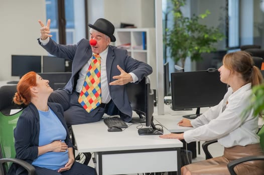 An elderly Caucasian man in a clown costume amuses two Caucasian women in the office