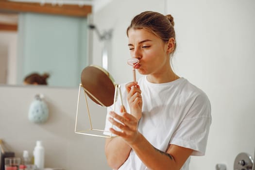 Pretty woman massaging face with jade roller and looking at round cosmetic mirror in bathroom