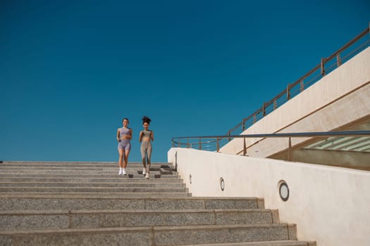 Two active female athlete friends in sportswear running on steps outdoors on a sunny day