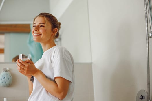 Smiling young woman drinking glass of water in the bathroom. Morning beauty routine