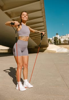 Young smiling woman makes fitness workouts with resistance band on morning city background