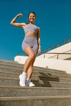 Smiling woman athlete raising arm and showing biceps while standing on modern buildings background