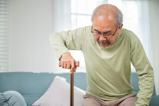 Elderly man suffering from knee pain ache holding handle of cane, senior old man typing to stand up from sofa alone with walking cane stick to walk at home, retirement medical healthcare concept