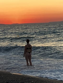 beautiful girl stands in the sea on the background of wonderful red sunset in Creta, Greece, 02 August 2023. young woman in black swimsuit staying by the waves of the sea water. Silhouette of a girl on the beach at sunset High quality photo