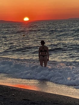 beautiful girl stands in the sea on the background of wonderful red sunset in Creta, Greece, 02 August 2023. young woman in black swimsuit staying by the waves of the sea water. Silhouette of a girl on the beach at sunset High quality photo