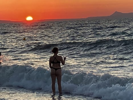 beautiful girl stands in the sea on the background of wonderful red sunset in Creta, Greece, 02 August 2023. young woman in black swimsuit staying by the waves of the sea water. Silhouette of a girl on the beach at sunset High quality photo