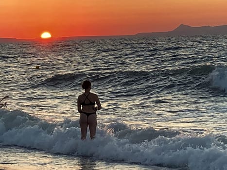 beautiful girl stands in the sea on the background of wonderful red sunset in Creta, Greece, 02 August 2023. young woman in black swimsuit staying by the waves of the sea water. Silhouette of a girl on the beach at sunset High quality photo