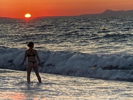 beautiful girl stands in the sea on the background of wonderful red sunset in Creta, Greece, 02 August 2023. young woman in black swimsuit staying by the waves of the sea water. Silhouette of a girl on the beach at sunset High quality photo