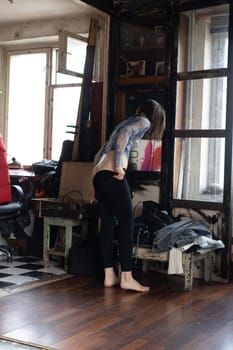 young beautiful woman posing nude in the studio, standing changing clothes near the window in black pants