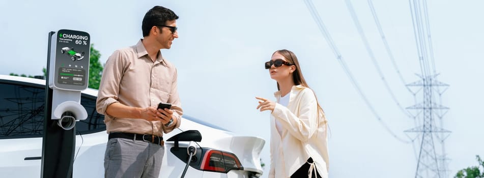 Couple pay for electricity with smartphone while recharge EV car battery at charging station connected to electrical power grid tower as industry for eco friendly car utilization. Panorama Expedient