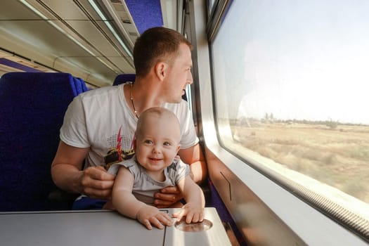 Young man - father, travelling with his little son by train, looking at the window. Dad with a laughing baby sitting at the table in the interior of the train