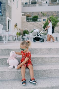 Little girl sits a pink toy soft rabbit nearby on the steps near an apartment building. High quality photo