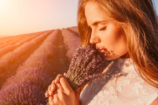 Close up portrait of young beautiful woman in a white dress and a hat is walking in the lavender field and smelling lavender bouquet.