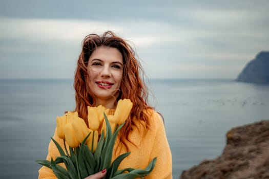 Portrait of a happy woman with hair flying in the wind against the backdrop of mountains and sea. Holding a bouquet of yellow tulips in her hands, wearing a yellow sweater.