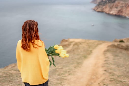 Rear view of a woman with long hair against a background of mountains and sea. Holding a bouquet of yellow tulips in her hands, wearing a yellow sweater.