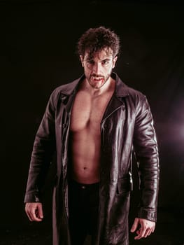 A man wearing a leather jacket on his muscular naked torso kneeling down on the ground in a casual yet attentive pose, his hands resting on his thighs.